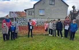 Grupa uczni&oacute;w z nauczycielami przy fladze Polski wykonanej z kolorowych wstążek.